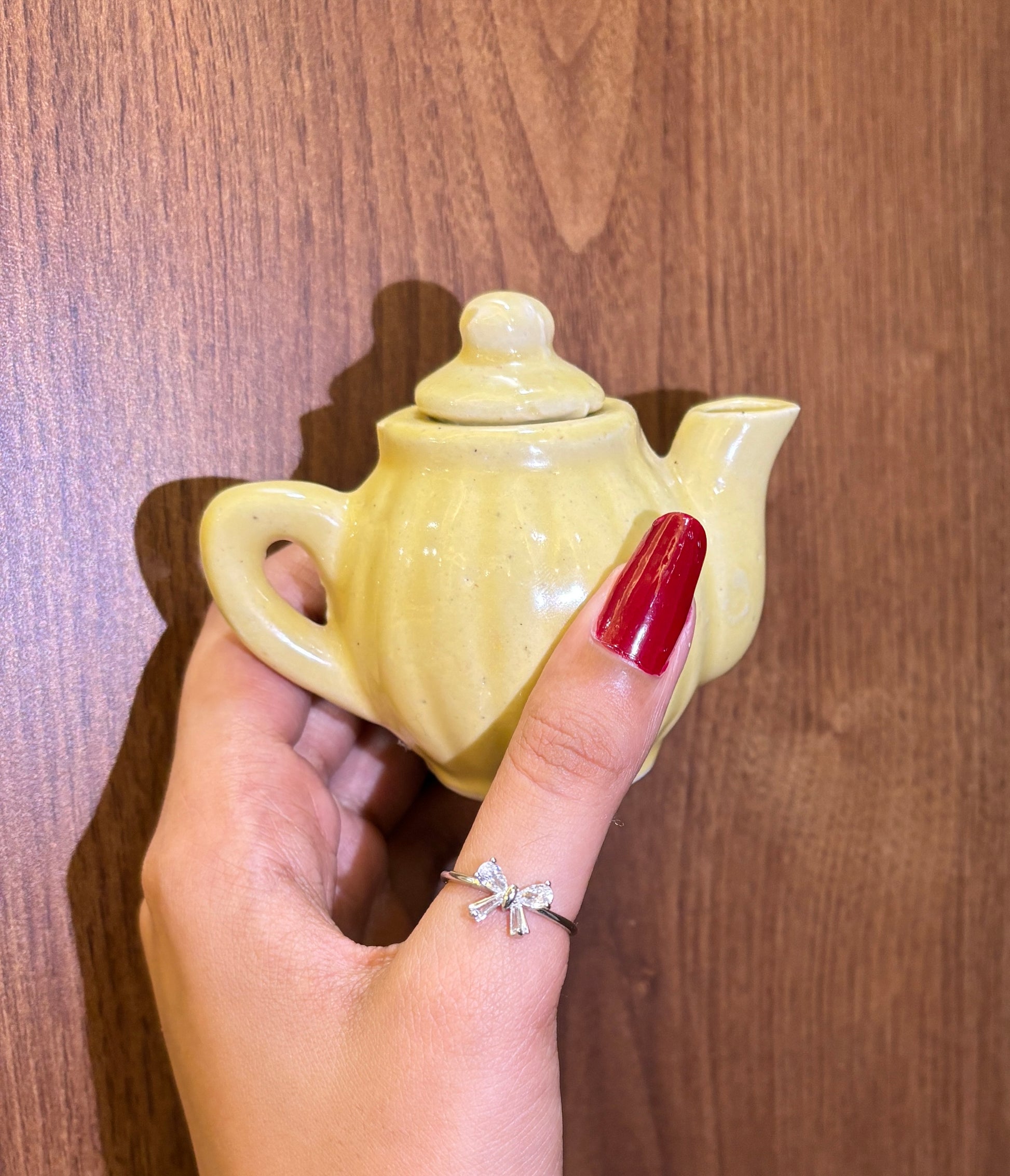 Hand wearing coquette bow ring holding a small yellow teapot against a wooden background