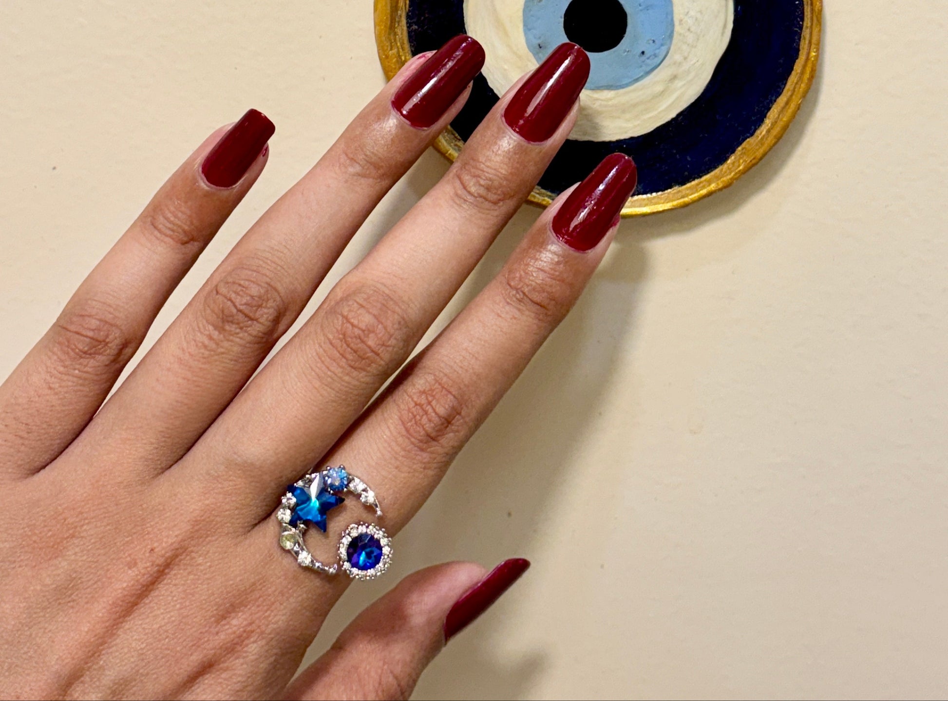 Hand with red nail polish wearing a blue and silver ring, holding a decorative evil eye charm.