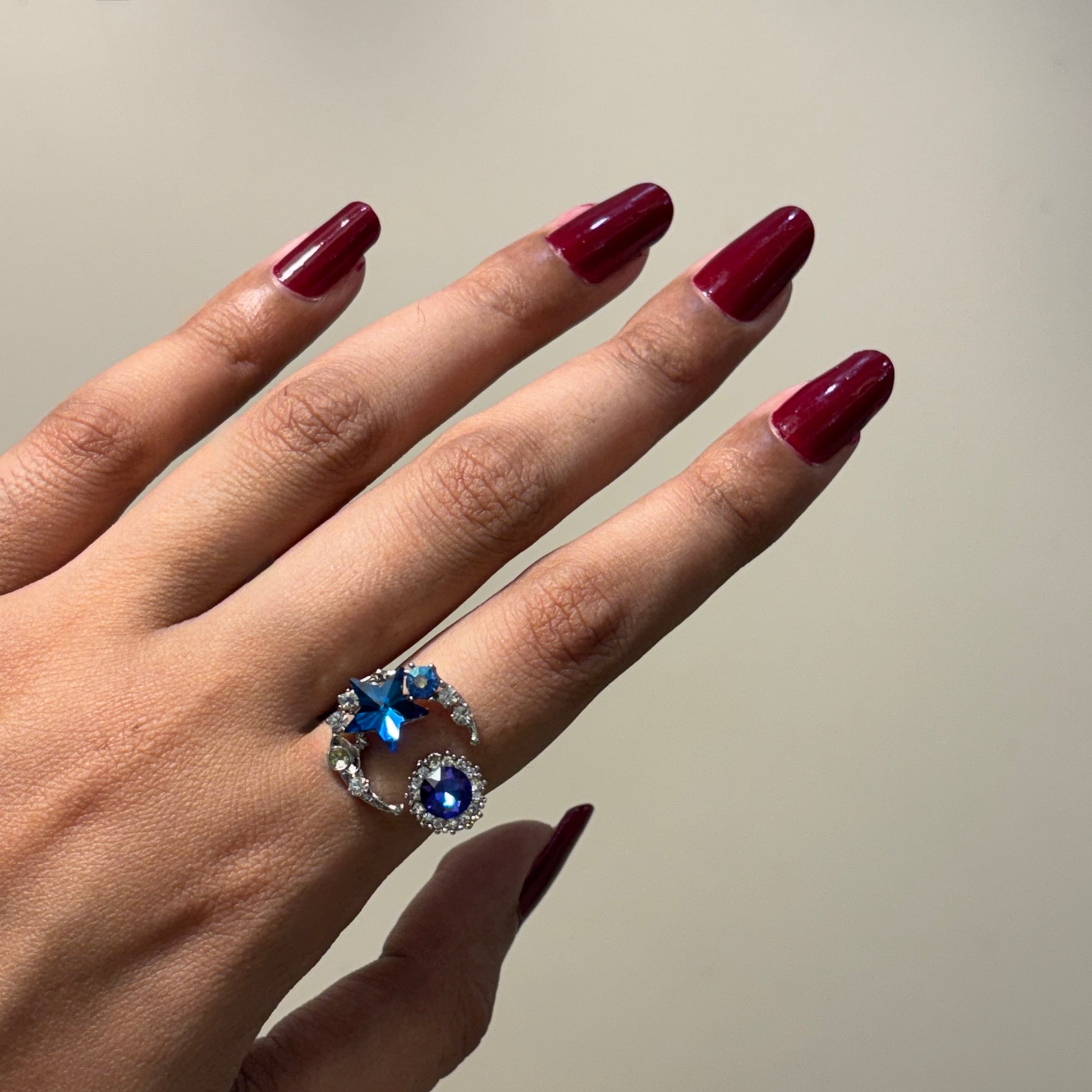 Hand with maroon nails wearing a silver ring with blue gemstones on a neutral background