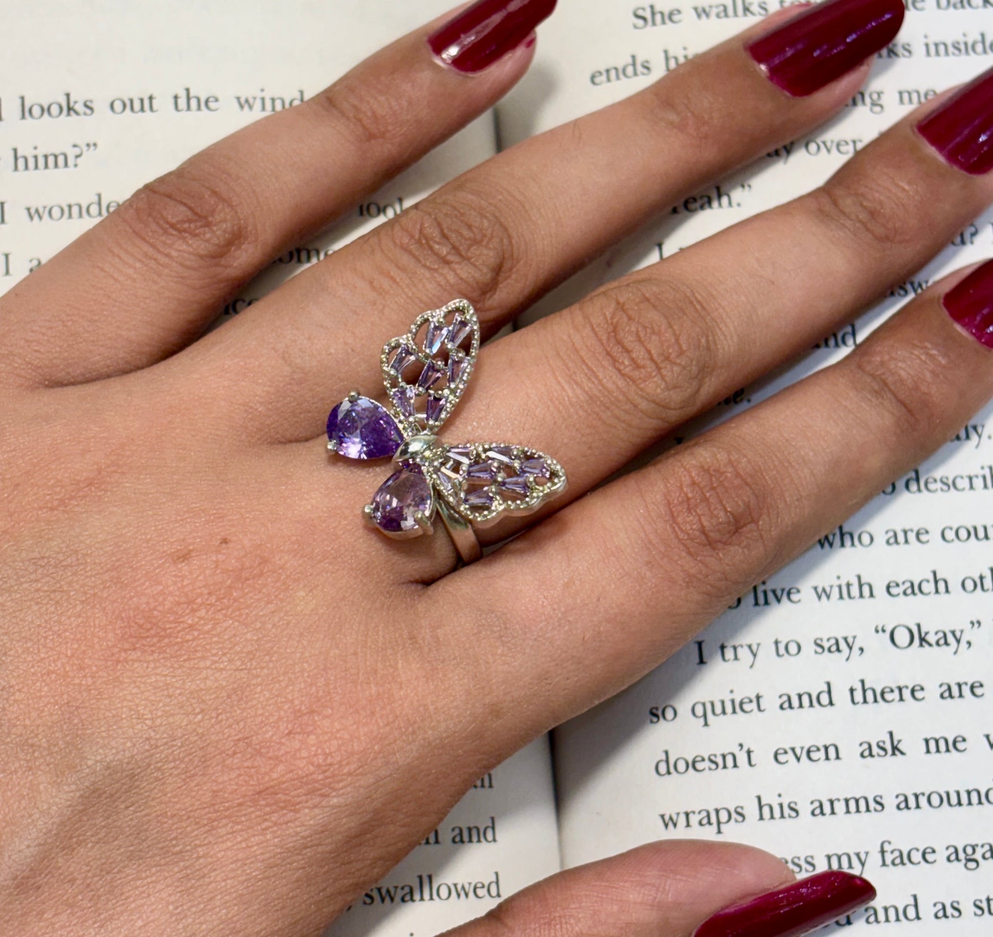 Butterfly-shaped ring with purple gemstones on a hand with red nail polish, against a book background.