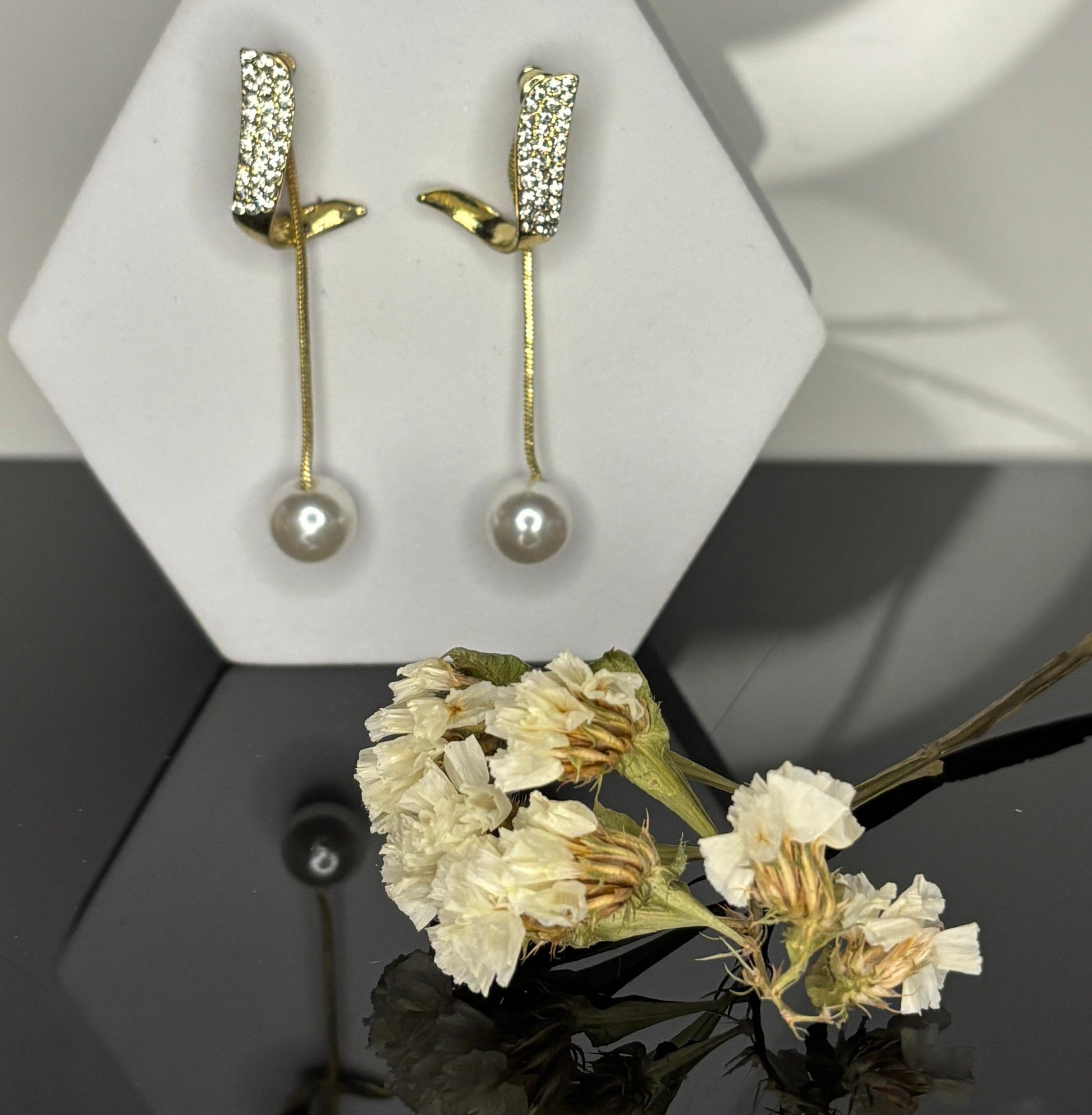 Gold earrings with pearl drops on a white stand, accompanied by dried flowers.