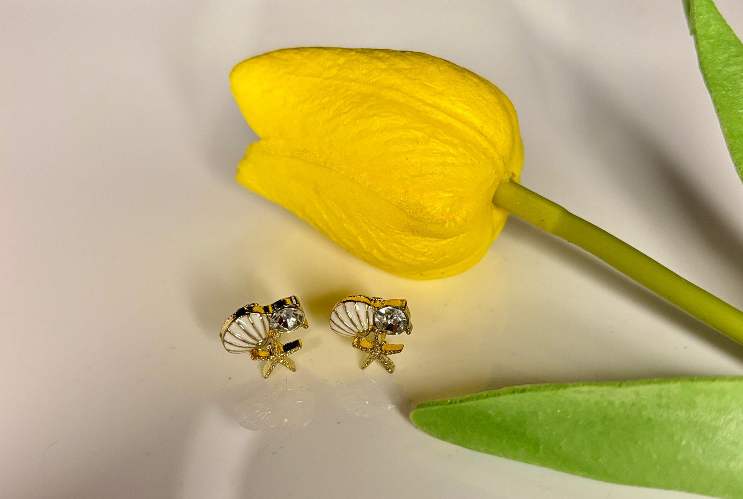 Gold shell-shaped earrings on a white surface with a yellow tulip in the background